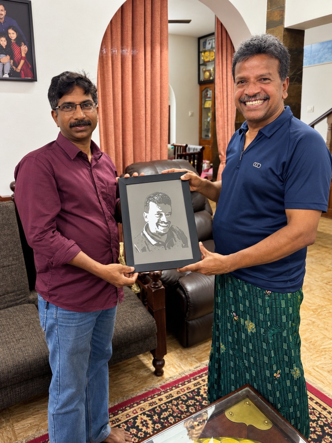 Two smiling men holding a framed paper-cut portrait together inside a home